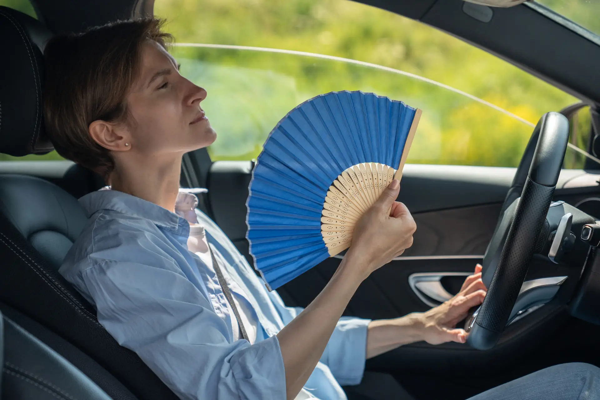 Overheated exhausted middle aged woman driver with hand fan suffering from heat in car. Hot weather